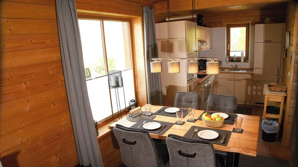 Un interior de madera con una mesa de comedor preparada para cuatro comensales, sillas, una zona de cocina con armarios y una ventana con cortinas.