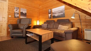 Interior acogedor de una cabaña con paredes de madera, un sofá, una silla, una mesa y una lámpara.