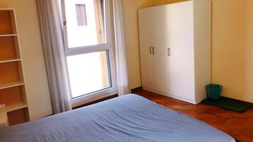A bedroom with a bed, a wardrobe, a window with curtains, and a trash bin.