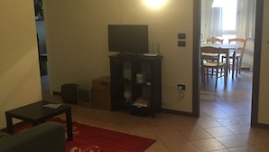 Een woonkamer met een bank, een salontafel, een tv-meubel en een rood vloerkleed. Er is een deur naar een eethoek met een tafel en stoelen.