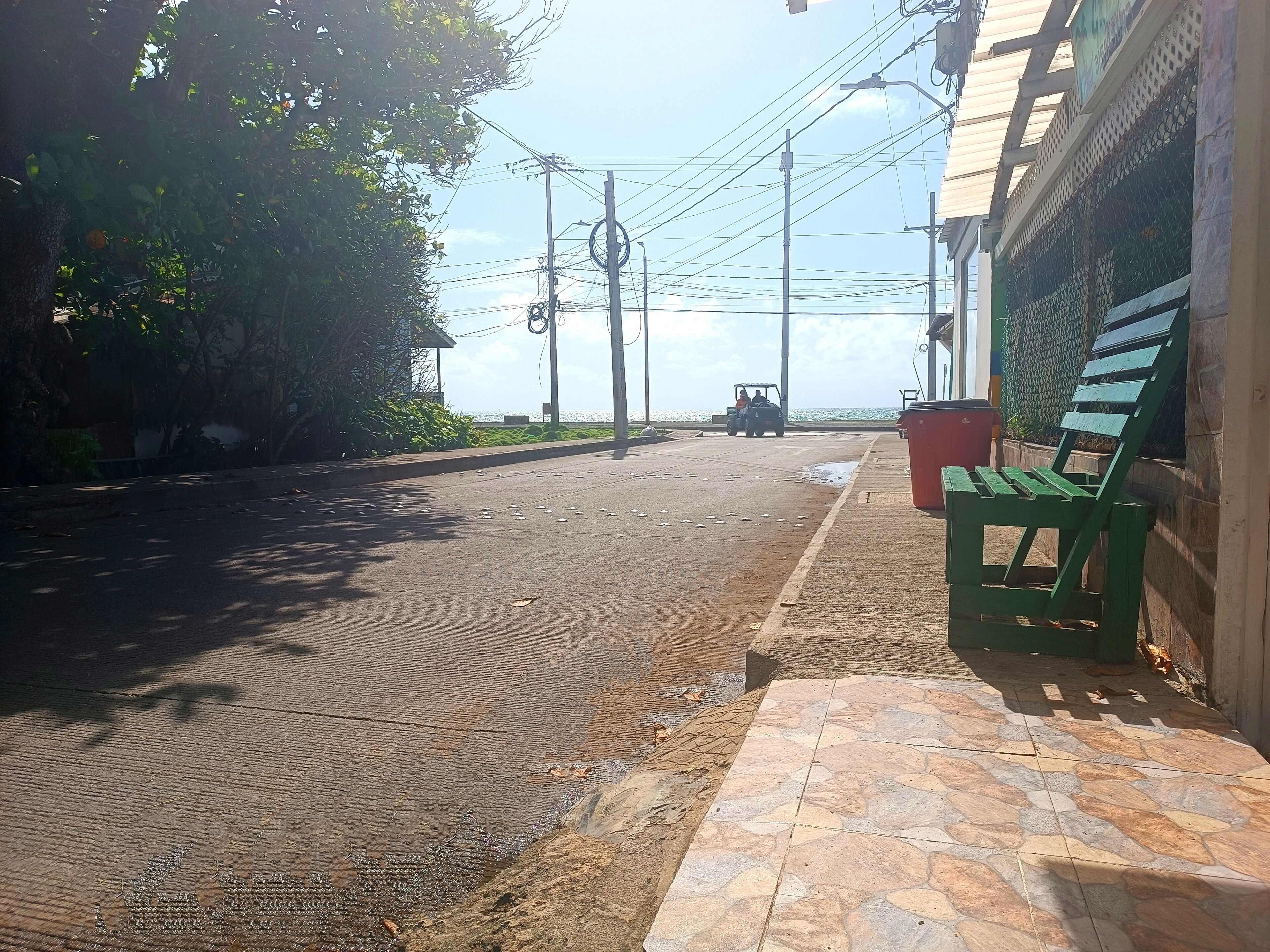 A paved street with a green bench, utility poles, and a distant view of the sea.