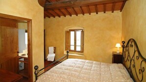 A bedroom with a wooden ceiling, a bed with a metal frame, a window with shutters, and a nightstand with a lamp. - Il Bellini - Tuscany Farmholiday (Castelfranco Piandisco)