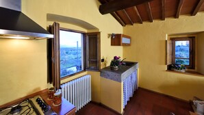 A compact kitchen with a gas stove, a window with a view, a radiator, and a small sink area with a basket of flowers. - Il Bellini - Tuscany Farmholiday (Castelfranco Piandisco)