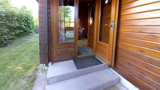 Entrada de una cabaña de madera con puerta de cristal, un porche pequeño y un interior visible con una silla y una lámpara.