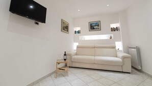 Una sala de estar con un televisor de pantalla plana montado en la pared, un sofá color beige, una pequeña mesita de madera con una botella de vino y una copa, y un radiador en el suelo.