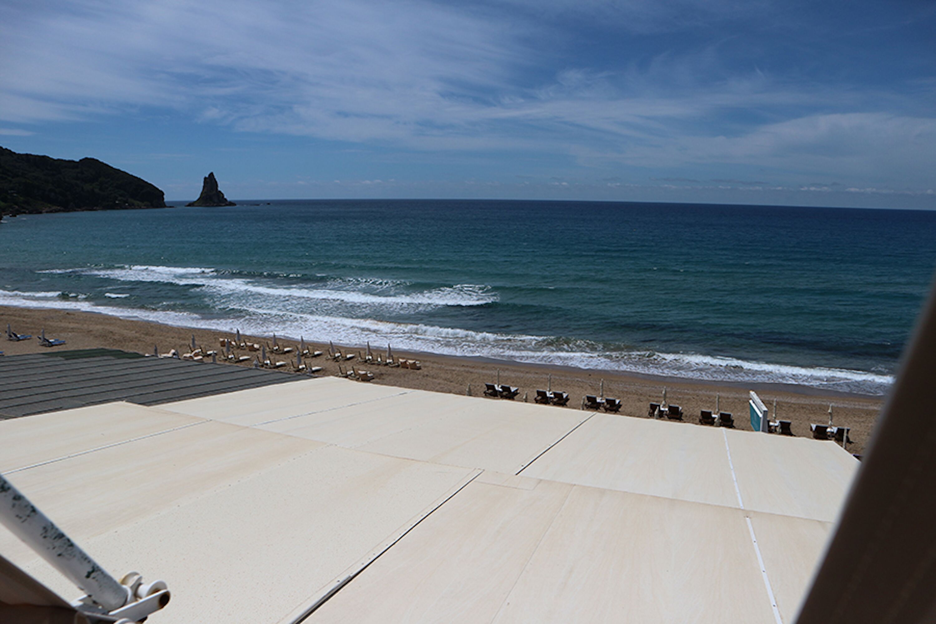 A beach with a rocky outcrop, a sandy shore with umbrellas, and a clear sky.