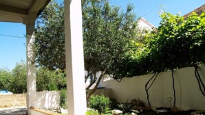 A covered patio area with a wooden deck, a white wall, and a garden with various plants and flowers. - Step Away From The Beach 1-bedroom Apartment Lolov (Preko)