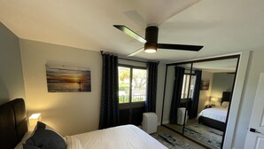 Makuuhuoneessa on suuri sänky, kattotuuletin, ikkuna, jossa on verhot, seinällä taulu ja peili, joka heijastaa huonetta.