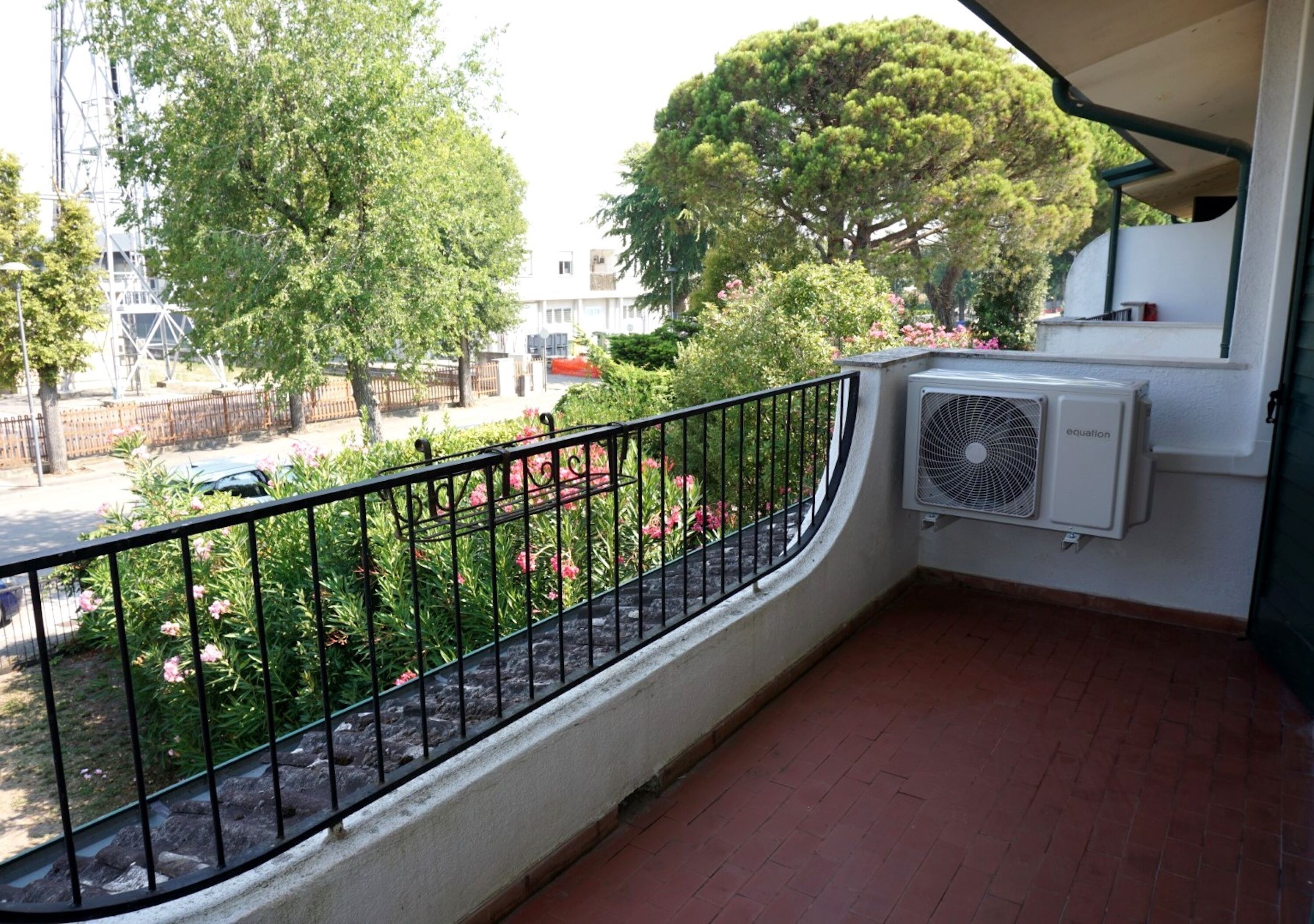 Un balcone con ringhiera in metallo, un piccolo condizionatore d'aria e vista su un giardino con alberi e fiori.