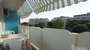 A balcony with a striped awning, a table with chairs, and a view of buildings and trees in the background. - Apartment 320mt From the sea (Caorle)
