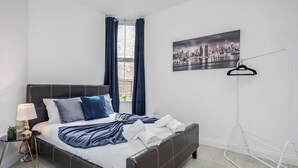 En pænt redt seng med puder, en sengetavlelampe, et bybillede og et vindue med gardiner.