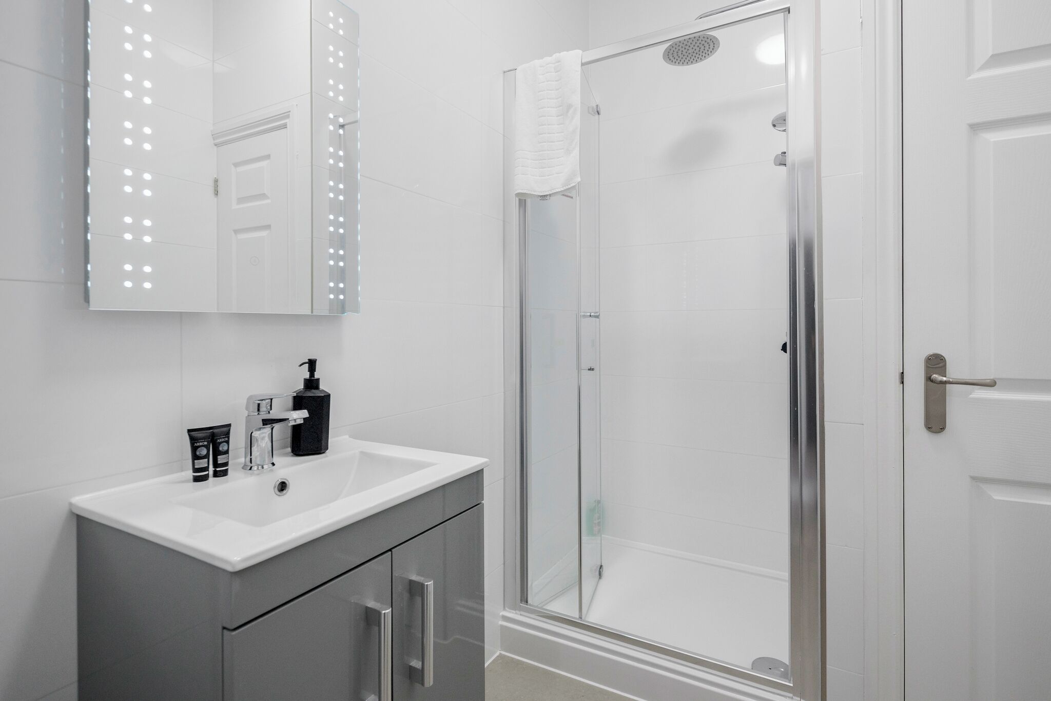 A bathroom with a white sink, a mirror with lights, a shower with a glass door, and a white door.