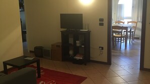 A living room with a sofa, a coffee table, a TV stand, and a red rug. There is a door leading to a dining area with a table and chairs.