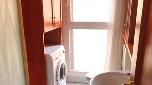 A compact bathroom with a washing machine, a sink with a gold faucet, and a window with a view of a building outside.