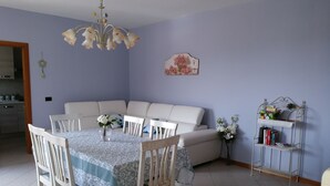 A dining area with a table set for four, surrounded by white chairs, a white sofa, and a decorative metal shelf with books and flowers. - Casa Annick Holiday Home (Barberino Tavarnelle)