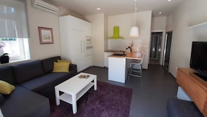 Uma sala de estar moderna com sofá, mesa de centro e televisão. Há também uma área de cozinha com mesa e cadeiras.