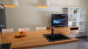Cozinha moderna com bancada de madeira, pia e uma fruteira. Há uma televisão sobre um suporte e uma estante branca ao fundo.