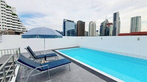 A rooftop swimming pool with lounge chairs and an umbrella, surrounded by high-rise buildings. - 7D - 3 Bedroom Downtown Apartment With Shared Pool (Bangkok)