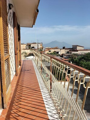 Uma varanda com piso de azulejos e grades de metal, com vista para uma área residencial com edifícios e uma montanha ao longe.
