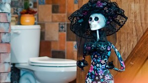 A skeleton figure in a floral dress and black lace hat standing in a bathroom with a tiled wall and a wooden door. - El Mezquite Hotel Mexicano Hab 1 la Catrina (Valle de Guadalupe)