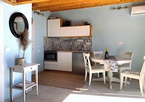 Cocina compacta con armarios blancos, microondas y lavavajillas. Hay una mesa de comedor con sillas y una mesita auxiliar con una planta.