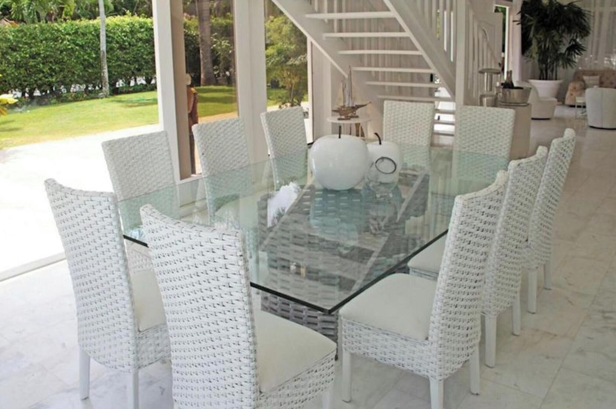 Una mesa con cubierta de vidrio rodeada de sillas de mimbre blanco en un espacio amplio y luminoso, con vista a un jardín y una escalera.