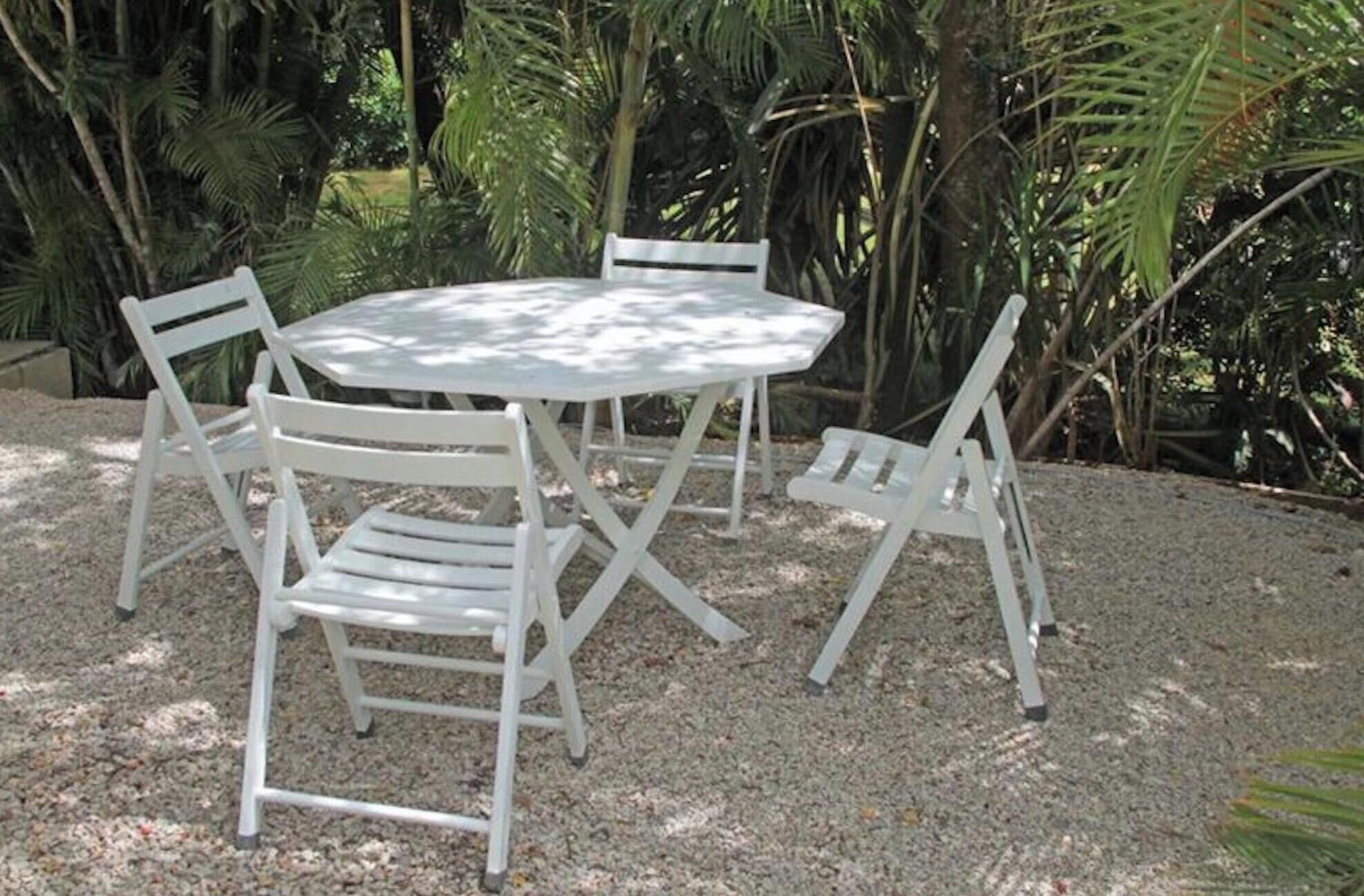 Mesa redonda al aire libre con cuatro sillas blancas, rodeada de grava y palmeras.