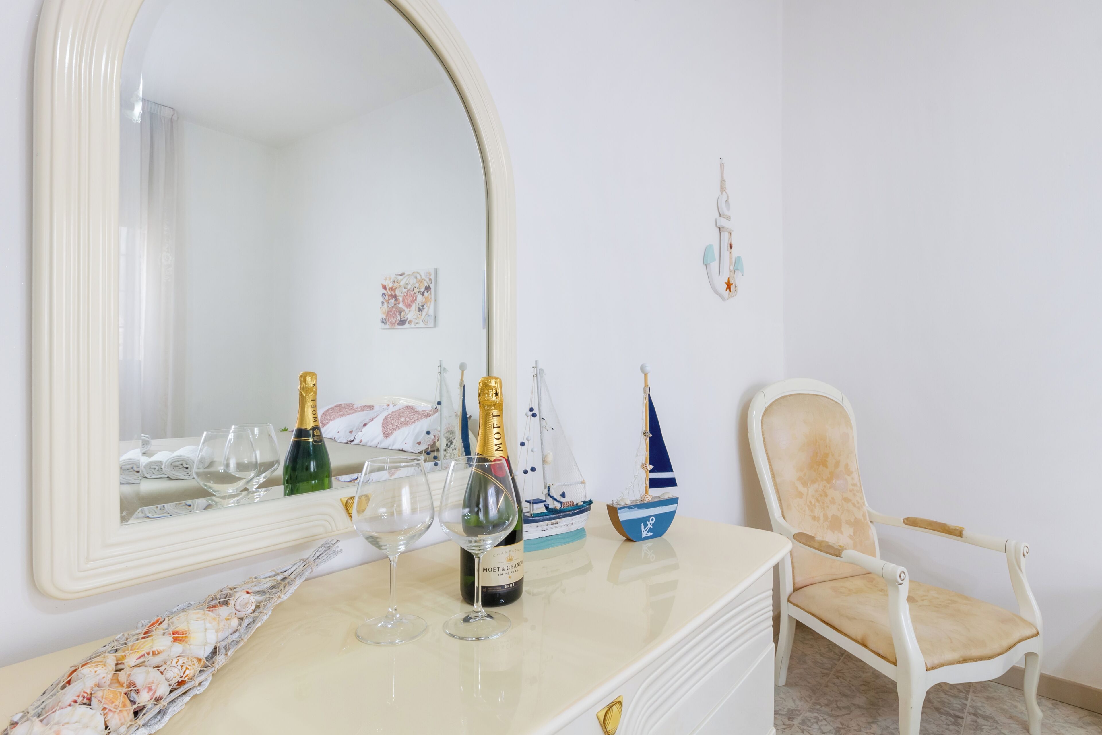 Une pièce comprenant une coiffeuse blanche, un miroir, une bouteille de champagne, deux verres à vin, un petit bateau de décoration et une chaise.