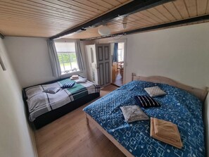 Huoneessa on kaksi sänkyä, puulattia, näkyviin jätetyt kattopalkit ja ovi, joka johtaa ulkotilaan.