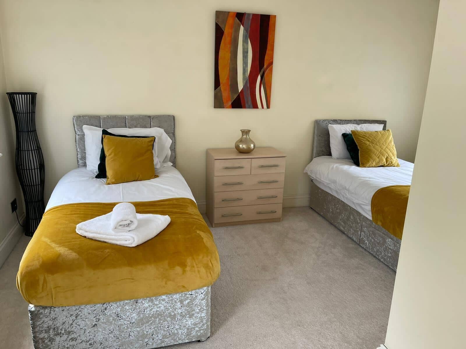 A hotel room with two beds, a bedside table, a lamp, and a painting on the wall.