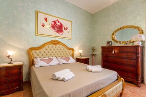 Un lit bien fait, avec une tête de lit, une table de chevet avec une lampe, une commode avec un miroir et une œuvre d’art florale encadrée accrochée au mur.