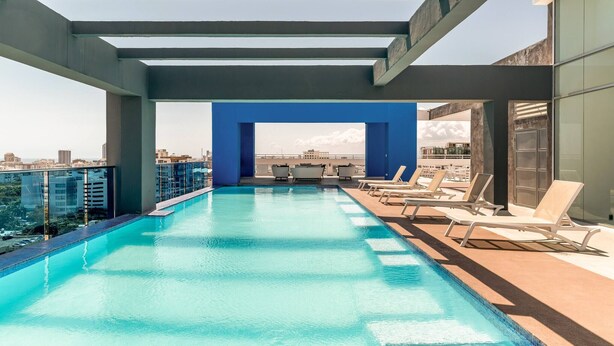 A rooftop infinity pool with a clear view of the city skyline, lounge chairs, and a blue architectural feature. - Apartamento S1404 - Downtown Santo Domingo (Santo Domingo)