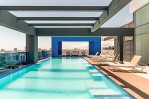 A rooftop infinity pool with a clear view of the city skyline, lounge chairs, and a blue architectural feature.