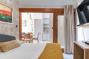 A modern hotel room with a bed, a TV, a desk, and a view of the kitchen and dining area.