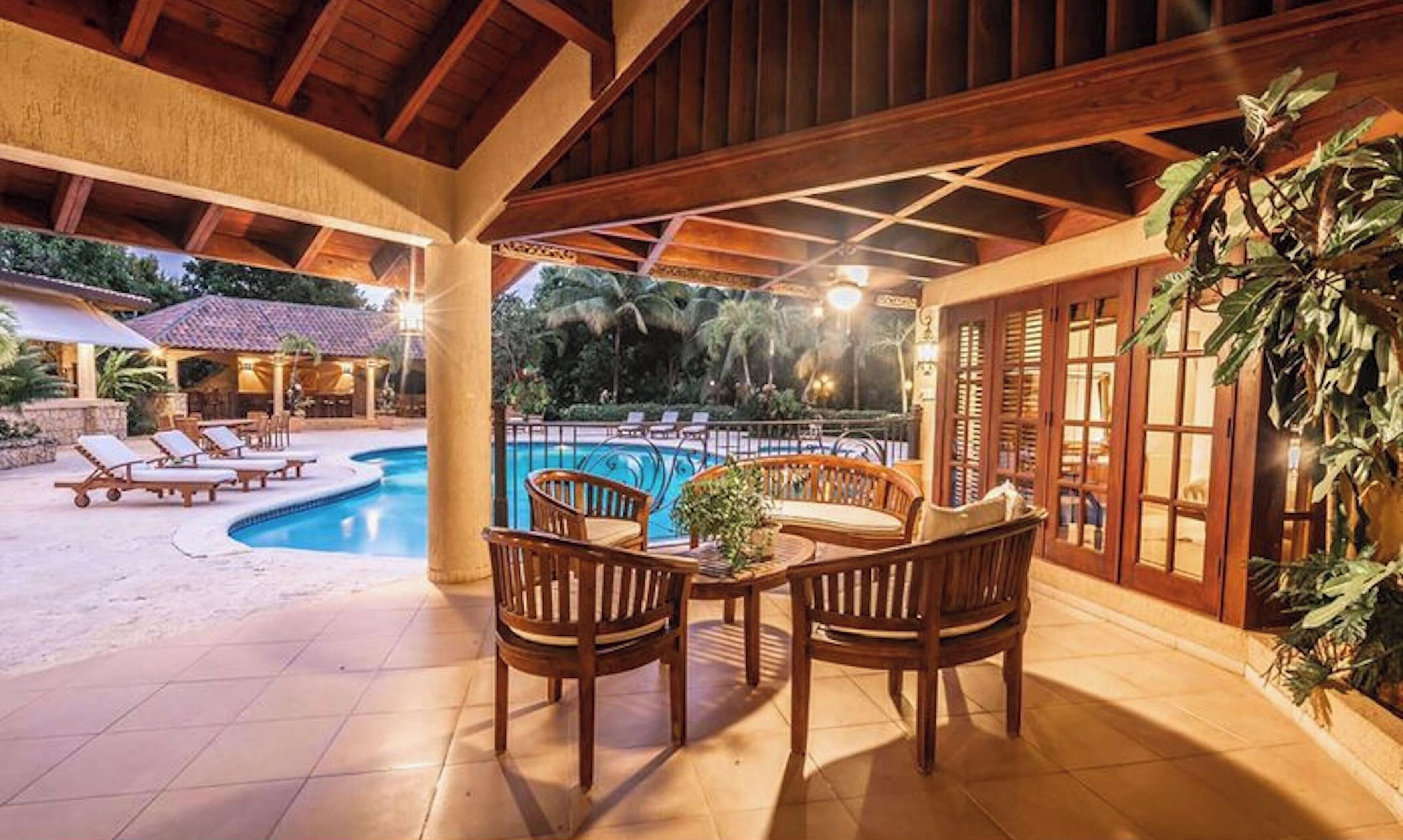 A covered outdoor area with a pool, lounge chairs, and wooden furniture.