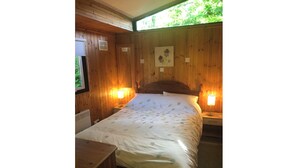 A wooden-paneled room with a bed, two bedside lamps, a radiator, and a window with a view of greenery. - Nuthatch Lodge is set in 24 Acres of Wood, Parkland Near the Village of Cenarth (Newcastle Emlyn)