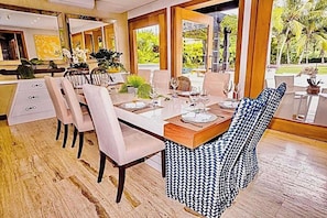 A dining area with a long table set for six, surrounded by chairs, with a view of greenery outside.