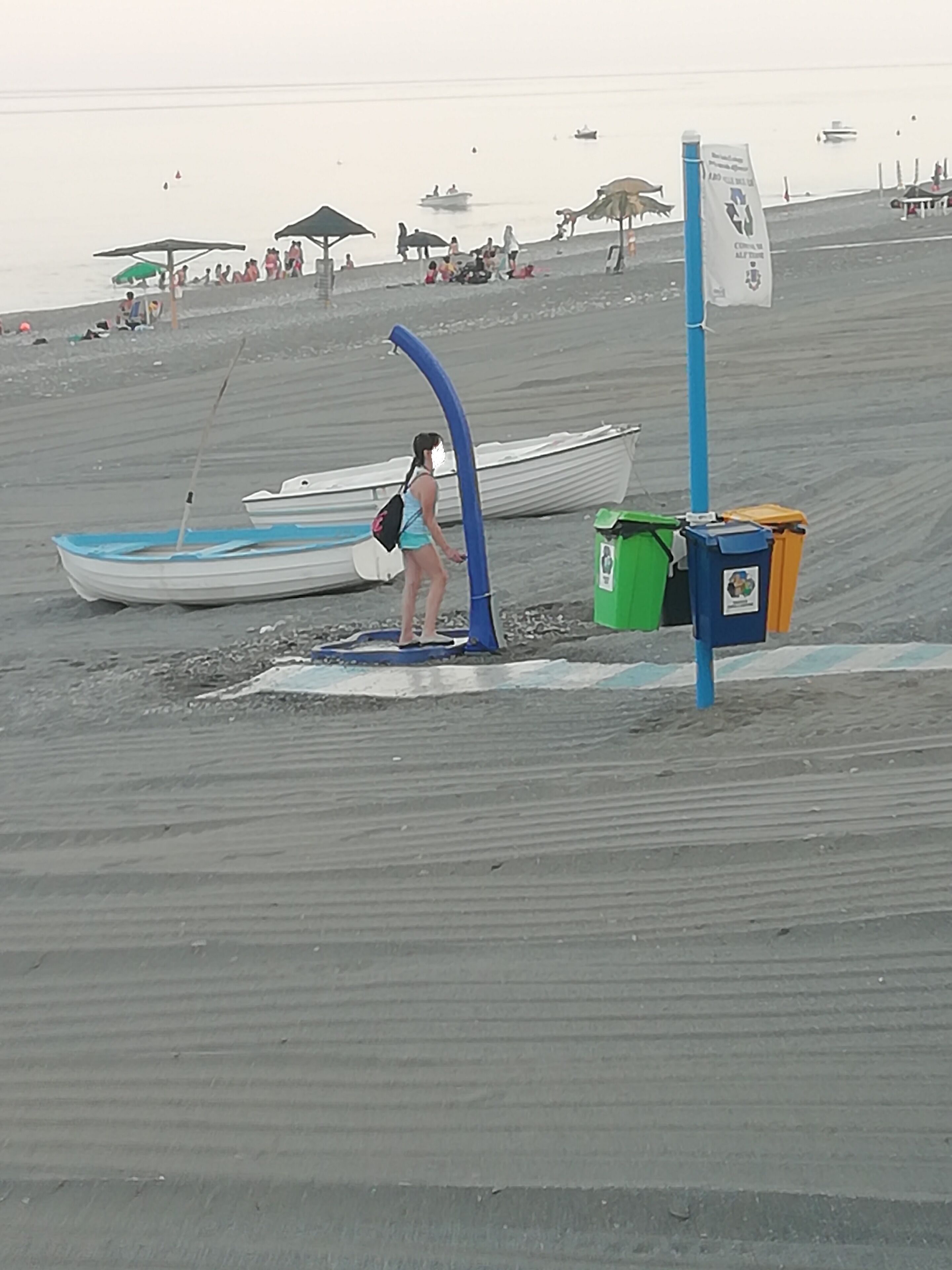 A beach with a woman standing near a blue structure, a boat, and trash bins.