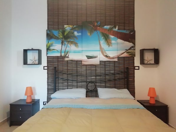 Une chambre à coucher comprenant un lit, deux tables de chevet, une tête de lit ornée d’une fresque sur le thème de la plage, et deux décorations représentant des étoiles de mer encadrées.