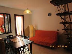 A dining area with a table and chairs, a sofa, a lamp, a clock, and a staircase.