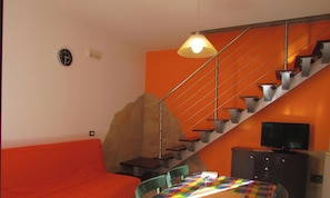 Un salon avec un canapé, une table à manger recouverte d’une nappe colorée, un escalier et un téléviseur posé sur un meuble.