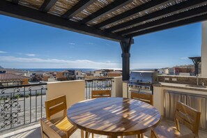 A covered outdoor patio with a round wooden table and chairs, a grill, and a view of a coastal town.