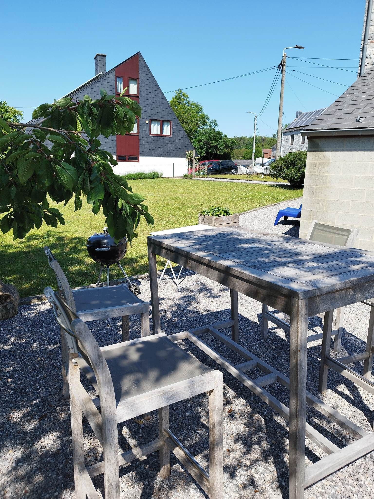 A wooden outdoor table and chairs set on gravel, with a house in the background.