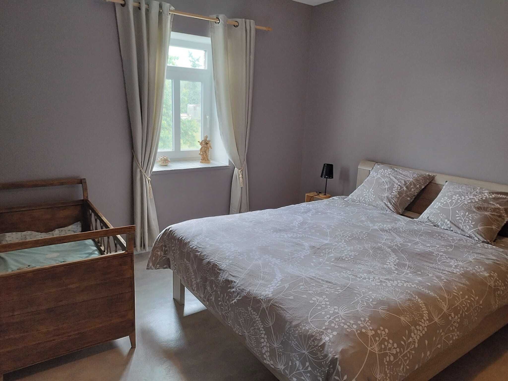 Quarto com estrutura de cama de madeira, uma cama grande com roupa de cama floral, mesa de cabeceira com abajur e janela com cortinas.