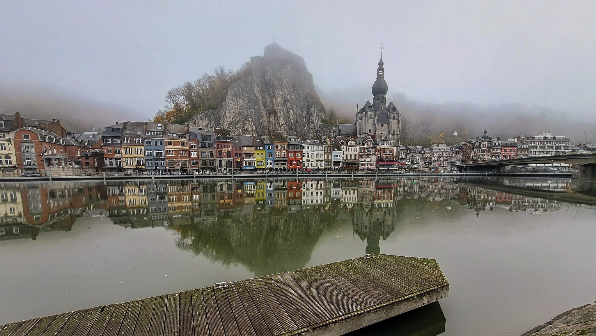 Un río tranquilo que refleja edificios coloridos, una iglesia destacada y una colina rocosa al fondo.