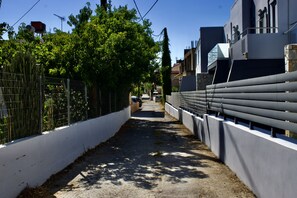 A narrow alleyway with a white wall on one side and a metal fence on the other, leading to a building with a modern design.