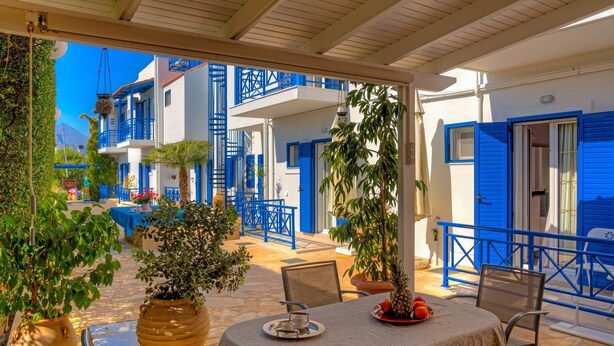 A covered outdoor seating area with a table set for two, surrounded by potted plants and a view of blue buildings. - Cozy Apartment Amoudara Heraklion Crera-4 (Malevizi)