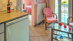A kitchen with a refrigerator, a wine bottle, and two glasses on the counter. There is a chair with a floral pattern and a table with red cups. A door leads to a balcony with a view of trees. - Cozy Apartment Amoudara Heraklion Crera-4 (Malevizi)