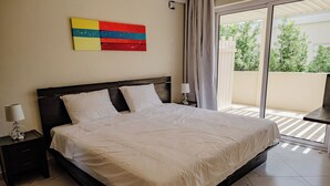 A hotel room with a large bed, a nightstand with a lamp, a colorful artwork on the wall, and a view of greenery through the balcony doors. - Charming 1-bedroom Apartment 2 km From Eagle Beach (Noord)