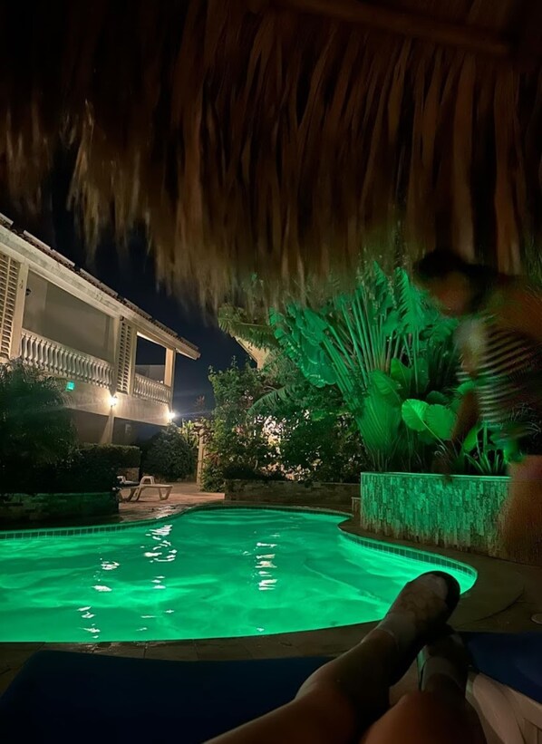 Ein Poolbereich mit einer strohgedeckten Struktur, ein Pool mit grüner Beleuchtung und ein Gebäude mit einem Balkon im Hintergrund.